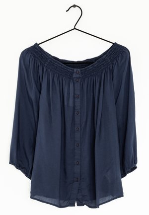 Granatowa bluzka z długimi rękawami, odsłaniająca ramiona, zapinana na guziki, zawieszona na czarnym drucianym wieszaku na białym tle.