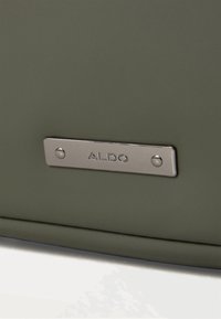 Bolso verde con una placa metálica plateada grabada con "ALDO", acentuado por una superficie lisa y texturada y un diseño minimalista.