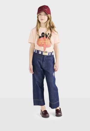 Camiseta de grafismo rosa claro con el texto "LOVE", pantalones de mezclilla de pierna ancha en azul, cinturón a rayas y gorra de béisbol burdeos. Mocasines marrones completan el atuendo.