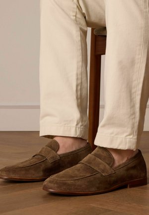 Brown suede loafers with a smooth texture and flat heel, featuring a stitched trim and a rounded toe, worn with light-colored trousers.