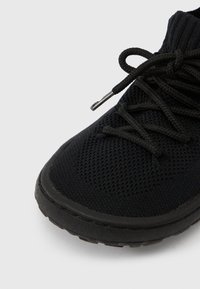 Μαύρο αθλητικό παπούτσι με υφασμάτινο επάνω μέρος με υφή, μαύρα επίπεδα κορδόνια και λαστιχένια σόλα με ελαφρύ ανάγλυφο σχέδιο.