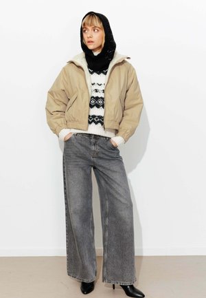 Veste beige courte avec poignets élastiques, pull noir et blanc à motifs, jean gris à jambes larges, et bottines noires. Fond blanc.