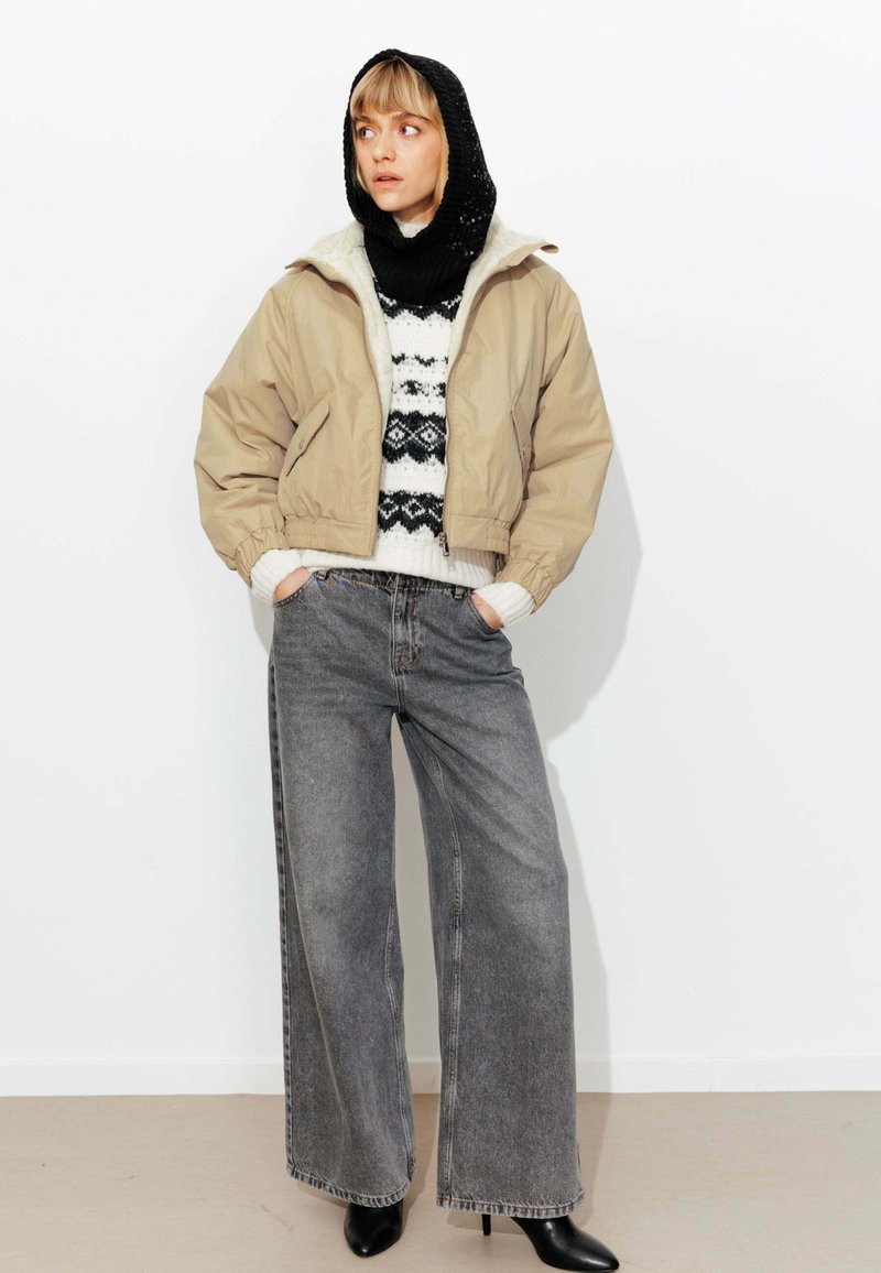 Veste beige courte avec poignets élastiques, pull noir et blanc à motifs, jean gris à jambes larges, et bottines noires. Fond blanc.