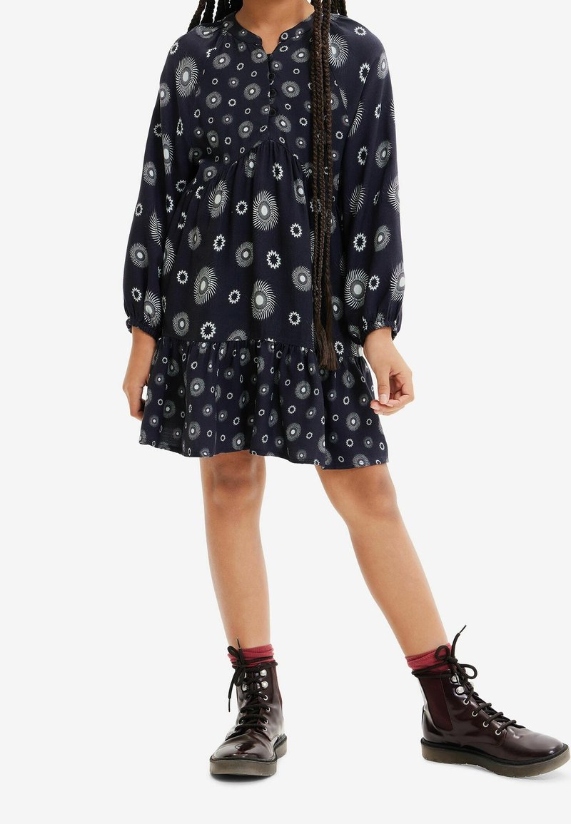 Robe bleu marine à motifs circulaires blancs, manches longues, encolure boutonnée, taille cintrée et ourlet à volants. Assortie à des bottes noires brillantes.