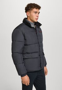 Giacca a vento in grigio scuro, con colletto alto, bottoni frontali, due ampie tasche e polsini a coste. Materiale testurizzato e isolato.