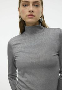 Haut gris à manches longues et col roulé avec un éclat métallique, présentant un design ajusté et une texture côtelée. Accentué par des boucles d'oreilles en argent.