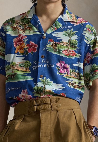Chemise bleue à manches courtes avec boutons, imprimée de motifs floraux et de joueurs de polo, rentrée dans un pantalon marron taille haute avec des détails de ceinture.