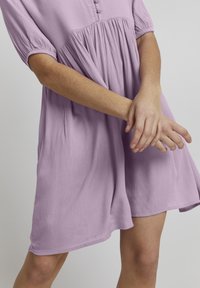 Lavendelfarbenes Kurzarmkleid aus strukturiertem Stoff, mit durchgehender Knopfleiste und einer lockeren, fließenden Silhouette, die bis zur Mitte des Oberschenkels reicht.