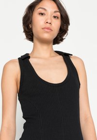 Top de alças preta, canelada, com decote redondo, apresentando costuras laterais curvadas e alças curtas cobertas de tecido nos ombros.