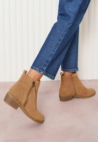 Botas de tobillo de ante color tan con cremallera lateral y tacón marrón texturizado, combinadas con jeans de mezclilla azul. Acabado liso y detalles de costura sutiles.