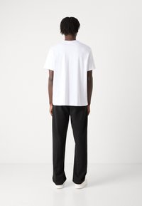 Hvid kortærmet t-shirt med runde halsudskæring, parret med sorte brede bukser og hvide sneakers, stående mod en ensfarvet baggrund.