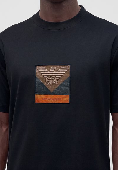 Μαύρο T-shirt με καφέ, μαύρο και πορτοκαλί λογότυπο Emporio Armani στο στήθος που δείχνει έναν αετό και τα γράμματα GA.