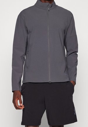 Homme portant une veste zippée gris foncé à manches longues et un short noir, debout devant un fond uni.