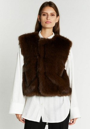 Femme portant une veste en fourrure marron foncé sur une chemise blanche à manches longues boutonnée et un pantalon noir, faisant face à l'avant avec une expression neutre.