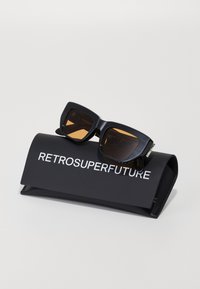 Mustad ristkülikukujulised prillid pruunide toonidega, puhkevad mustal ümbrisel, millel on valge tekst "RETROSUPERFUTURE".