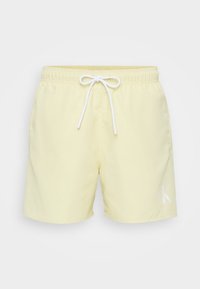 Shorts de bain jaune clair en tissu lisse, avec une taille élastique et un cordon de serrage blanc, ainsi qu'un logo blanc sur la cuisse gauche.