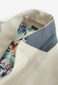 Veste en lin beige avec un col en feutre gris. À l'intérieur, elle présente une doublure à imprimé floral dans des teintes pastel. Des coutures soignées accentuent les bords.