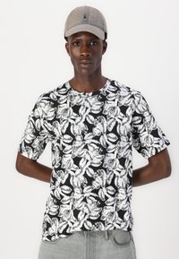 Sort t-shirt med et hvidt blomsterdesign, korte ærmer og rund halsudskæring. Modellen bærer en lysegrå kasket.