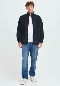 Svart fleecejakke med høy krage og frontglidelås, båret over en hvit T-skjorte, paret med blå jeans og lyse sneakers.