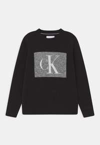 Černý svetr s kulatým výstřihem, na přední straně je šedý pletený obdélník s bílými písmeny "CK". Žebrované manžety a lem.