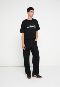 Μαύρο υπερμεγέθες μπλουζάκι με λευκό γραφικό τύπωμα, συνδυασμένο με φαρδιά μαύρα παντελόνια και μαύρα αθλητικά παπούτσια με λευκή και καφέ σόλα.