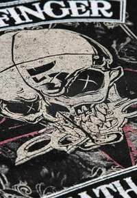 Černá tkanina s vytištěným designem lebky s metalickými akcenty, složitou linkovou prací a červenými detaily. Text v horní části zní "FINGER."