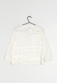 Hvid letvægtsbluse med 3/4 ærmer, gennemsigtigt stof, vandrette crochetsyninger og blondekant forneden. Hænger fra en sort krog.