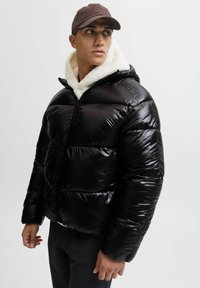 Giovane uomo che indossa una giacca a vento nera lucida sopra un hoodie bianco in pile e un cappello marrone, che guarda di lato su uno sfondo semplice.