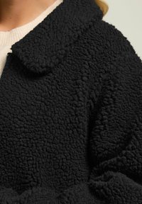 Mustapintainen teksturoitu takki, jossa on pehmeä ja pörröinen ulkonäkö. Varustettu pyöreällä kauluksella ja vetoketjulla, mikä tuo rentoa ilmettä.