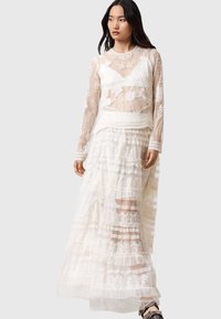 Weißes Spitzenkleid mit langen Ärmeln, das geschichtete Rüschen und Blumenstickerei bietet. Durchscheinende Textur und aufwendige Details im gesamten Kleid.