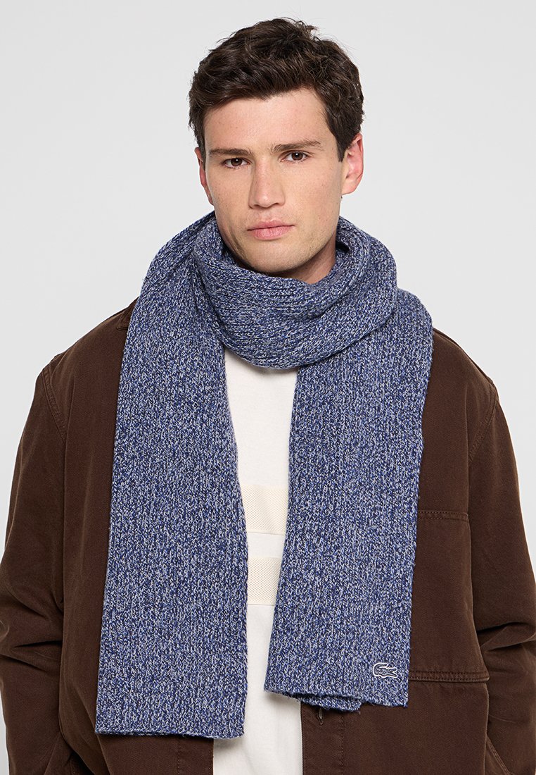 Écharpe tricotée bleu marine avec un motif texturé, agrémentée d'un petit logo. Portée lâchement autour du cou, associée à une chemise légère et un manteau.