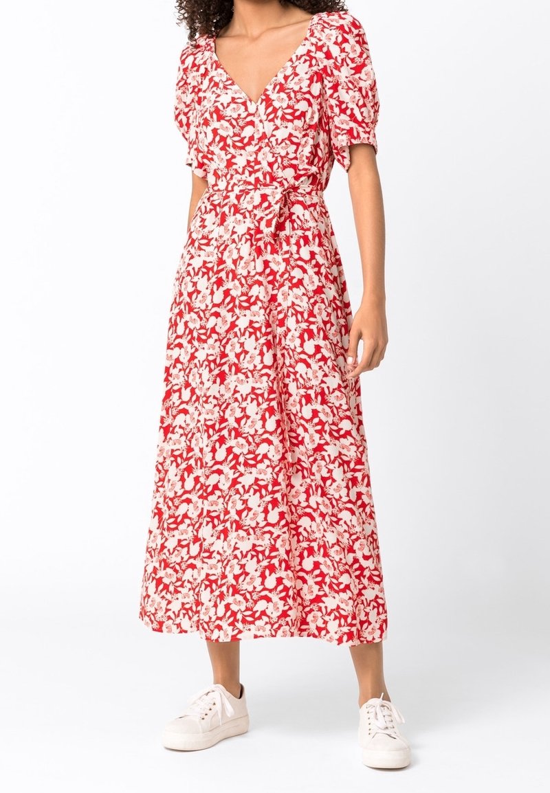Femme portant une robe portefeuille midi à fleurs rouges et blanches avec des manches bouffantes et des baskets blanches, debout devant un fond uni.