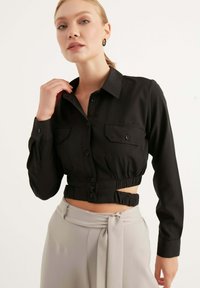 Camicia nera, cropped, con bottoni e maniche lunghe, caratterizzata da due tasche sul petto e una vita arricciata. Tessuto liscio con una finitura elegante.