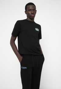 Črna bombažna majica s kratkimi rokavi z majhnim natisnjenim logotipom na prsih, kombinirana s črnimi trenirkami, ki imajo modro natisnjeno podrobnost ob strani.