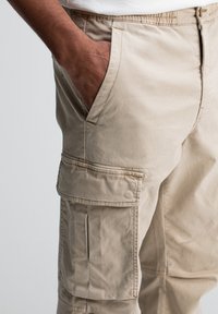 Donkere hand rustend in de voorzak van beige cargopantalons met zichtbare zijflapzak en knoopsluiting.