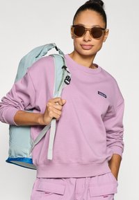 Lys lilla genser i crewneck-stil, brukt med mintgrønn ryggsekk med grå stropper og blå detaljer. Runde, mørke solbriller.