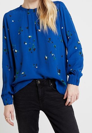 Femme portant un chemisier bleu à manches longues avec de petits imprimés floraux, rentré dans un jean noir taille haute, montrant des bagues aux doigts.