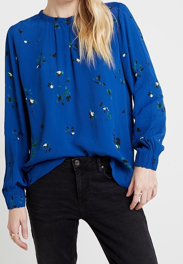 Femme portant un chemisier bleu à manches longues avec de petits imprimés floraux, rentré dans un jean noir taille haute, montrant des bagues aux doigts.
