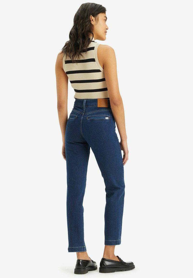 Jean taille haute bleu foncé avec une coupe droite, cinq poches et un patch en cuir beige à l'arrière de la taille. Des mocassins noirs sont portés.