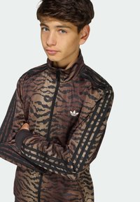 Hnedo-čierna vzorovaná bunda na zips s vysokým golierom, s tromi čiernymi pruhmi na každom ramene a bielym logom na hrudi.