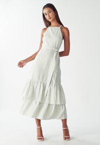 Dryžuota maxi suknelė šviesiai žalia ir balta, su kaklo apykakle, diržo juosmeniu ir daugiakampėmis volanėlėmis apatinėje dalyje. Lygus audinio tekstūra.