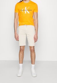 Oranžové tričko s kulatým výstřihem a bílým logem "Calvin Klein Jeans"; krémové teplákové kraťasy s tkaničkou; bílé tenisky a ponožky.