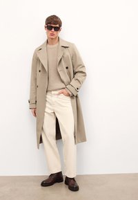 Beżowy płaszcz trench, jasnobrązowy sweter z dzianiny, kremowe spodnie i ciemnobrązowe buty. Płaszcz ma podwójne guziki i klasyczny fason.