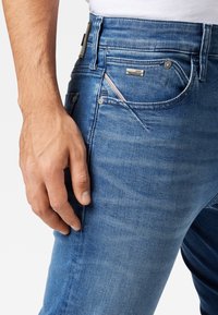 Manlig hand vilande nära framfickan på blå denimjeans med synliga sömmar, nitar och en liten logotavla.