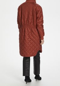 Pikowana długa kurtka w kolorze rdzy z wcięciem w talii, prążkowanymi mankietami i wysokim kołnierzem. Noszona na dopasowane czarne spodnie i botki na obcasie.