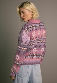 Neulepusero vaaleanpunaisissa ja violetissa sävyissä, jossa on geometrisia kuvioita ja lumihiutaleita. Ribbireunus helmassa ja hihansuissa, rento malli. Yhdistetty vaaleisiin farkkuihin.