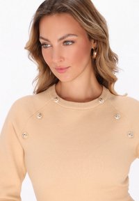 Beige Strickpullover mit Rundhalsausschnitt. Verfügt über dekorative Knöpfe an der Schulter. Glatte Textur, taillierte Passform und nahtlose Akzente.