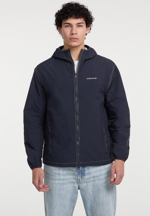 Giacca con cappuccio blu navy con chiusura a zip e dettagli di cucitura bianci. Presenta tasche laterali e una vestibilità rilassata. Il materiale appare leggero e liscio.