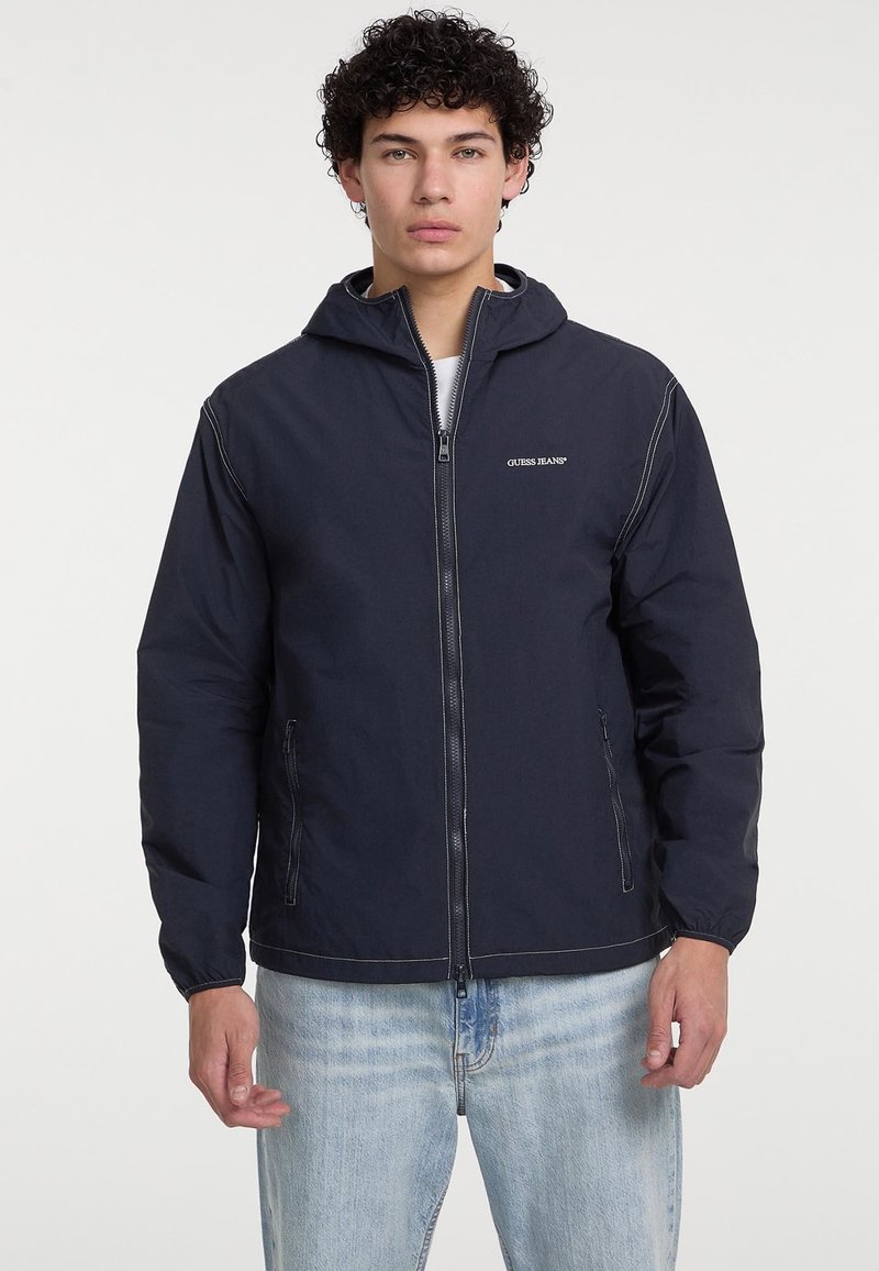 Giacca con cappuccio blu navy con chiusura a zip e dettagli di cucitura bianci. Presenta tasche laterali e una vestibilità rilassata. Il materiale appare leggero e liscio.
