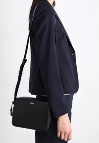 Person som bär en marinblå kavaj och har en svart rektangulär crossbody-väska med "BOSS"-logotyp på ett tunt justerbart rem.
